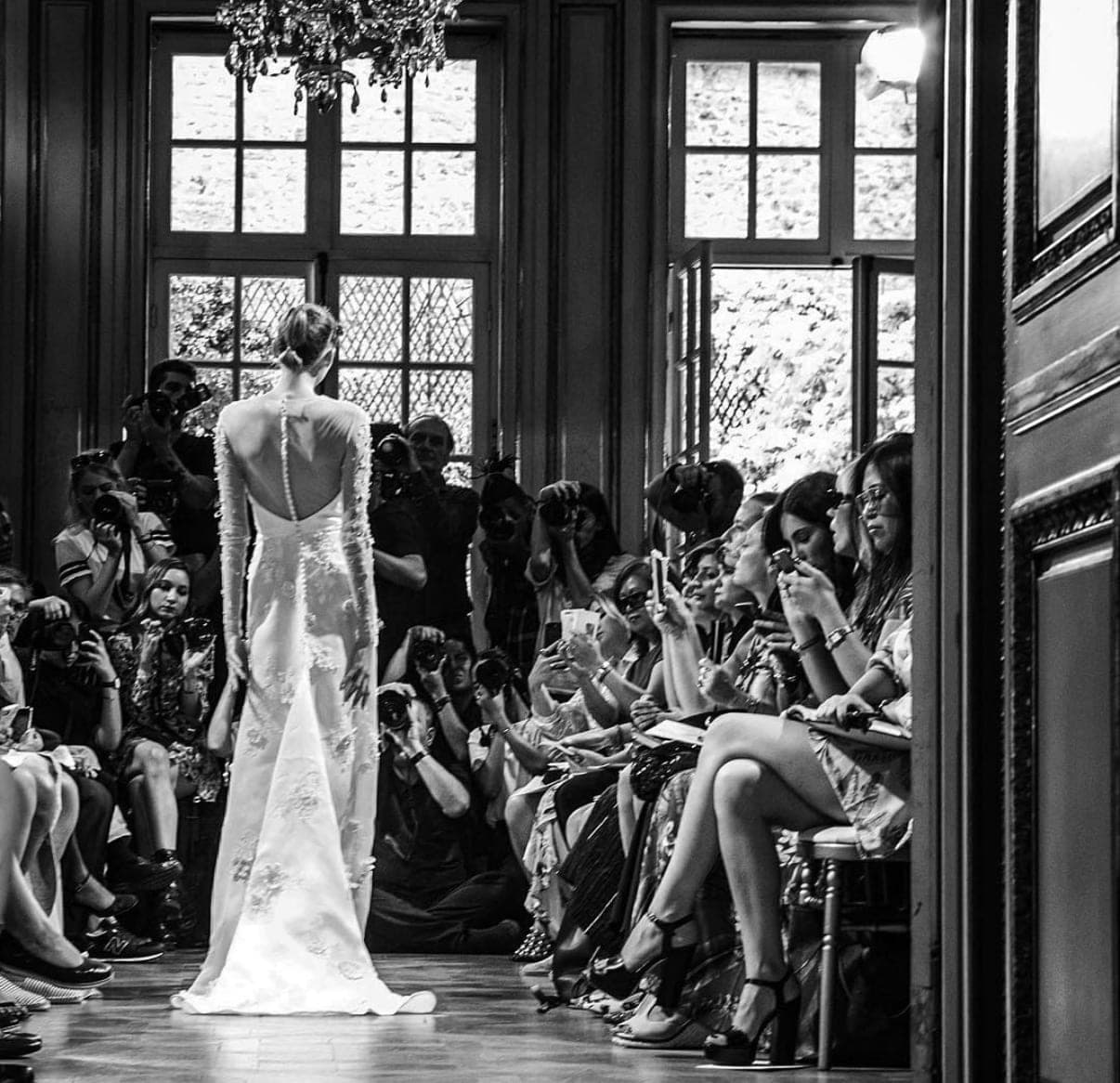 Backstage at a fashion show — a model in a gown surrounded by photographers, in black and white